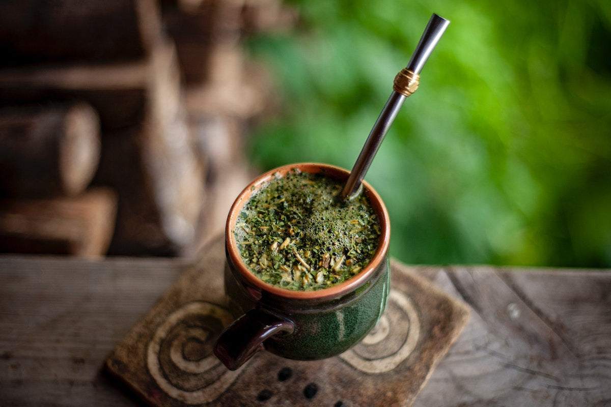 Eine ansprechende Szene mit einer Tasse gefüllt mit Mate Tee im Vordergrund, umgeben von einem beruhigenden, grünen Hintergrund, der reich an natürlichen Pflanzen ist, und eine Atmosphäre der Ruhe und Entspannung vermittelt.